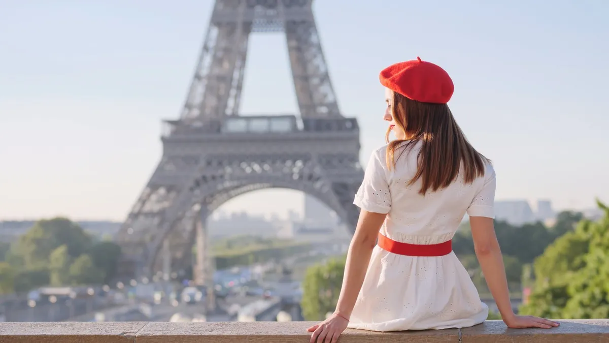 A woman in a red beret and white dress sits with her back to the camera looking at the Eiffel Tower reminiscent of Emily in Paris